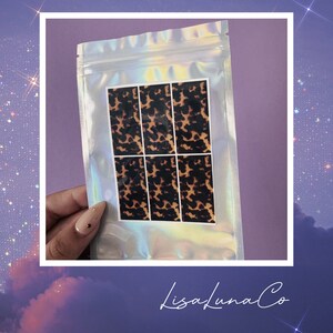 May include: A clear plastic bag with a sheet of six brown and black patterned nail decals inside. The bag has a holographic iridescent finish. The text "LisaLunaCo" is printed on the bottom of the image.