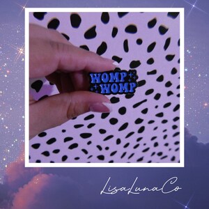 May include: A small, rectangular enamel pin featuring a black background with the text "WOMP WOMP" in a bold, bright blue font. The design includes several small, four-pointed star accents scattered around the lettering, creating a decorative effect.