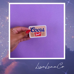 May include: A rectangular embroidered patch featuring a tan background with the blue text Coors above a red banner containing the white text Banquet. The patch has a white border and includes small gold lion illustrations on the left and right sides.