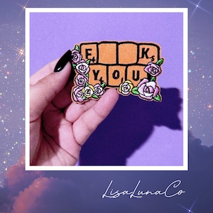 May include: A colorful embroidered patch with the words "F*ck You" spelled out in orange and yellow letters, surrounded by pink and yellow flowers. The patch is held in a hand against a purple background.