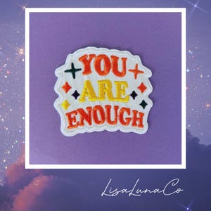 May include: White embroidered patch with the text "You Are Enough" in orange, yellow, and green lettering. The patch has a white border and a plus sign on either side of the word "You".