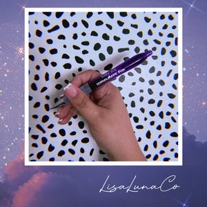 May include: Purple pen with white lettering that says "Take me somewhere fun". The pen is held in a hand against a white background with black polka dots.