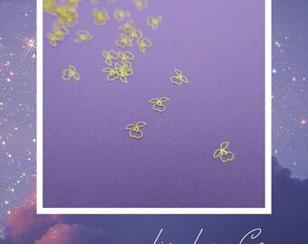 May include: A scattering of gold metal flower charms on a purple background. The charms are small and delicate, with a simple design. The background is a solid purple color.