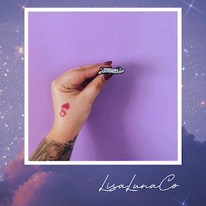 May include: A black enamel pin with the words "PROBABLY OVERSTIMULATED" in white lettering. The pin is held against a purple background. The image also includes a tattoo of a playing card and the text "LisaLunaCo".