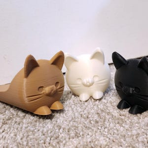 May include: Three cat-shaped doorstops in brown, white, and black. Each doorstop has a flat base and a rounded cat head with small ears, a nose, and whiskers. The doorstops are made of a 3D-printed material.