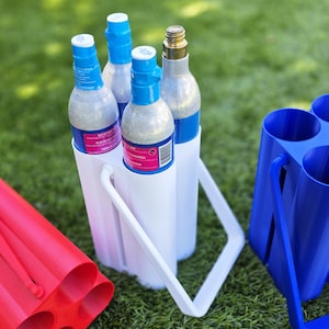 May include: A white plastic carrier holds four silver and blue soda bottles. A blue plastic carrier with four cylindrical openings sits nearby. A red plastic carrier with four openings is in the foreground. All are on green grass.