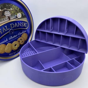 May include: A blue and gold Royal Dansk Danish Butter Cookies tin next to a purple, semi-circular storage container with multiple compartments. The tin has an image of a building and the words "Royal Dansk" and "Danish Butter Cookies". The container is open.