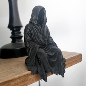 May include: A small, black, three-dimensional figurine of a hooded figure in a seated position. The figure wears a long, draped robe with visible folds and a deep hood that obscures the face, creating a dark, monochromatic appearance.
