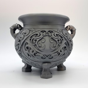 May include: A matte dark gray cauldron featuring intricate raised scrollwork and filigree patterns across its rounded body. The vessel is supported by three sturdy feet and includes two curved side handles, with a wide, smooth, circular rim.