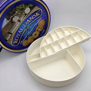 May include: A blue and gold Royal Dansk Danish Butter Cookies tin next to a white, multi-compartment storage container. The tin features an image of cookies and a landscape. The storage container is open, revealing several sections.