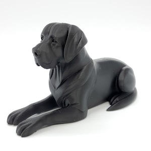 May include: A black Labrador Retriever statue in a resting pose. The dog is lying down with its front paws extended. The statue is made of a solid, dark material and has a smooth finish. The dog's features, including its ears and snout, are clearly defined.
