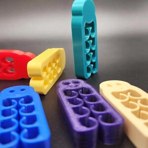 May include: A collection of colorful, abstract, 3D-printed objects in red, yellow, blue, purple, and teal. Each object has a unique shape with multiple curved cutouts, arranged on a dark surface.