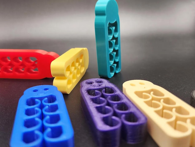 May include: A collection of colorful, abstract, 3D-printed objects in red, yellow, blue, purple, and teal. Each object has a unique shape with multiple curved cutouts, arranged on a dark surface.