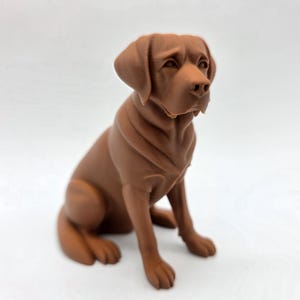 May include: A brown dog figurine in a seated position, with a focused gaze. The sculpture has detailed features, including the dog's ears, snout, and paws. The dog is a solid color and sits on a white surface.