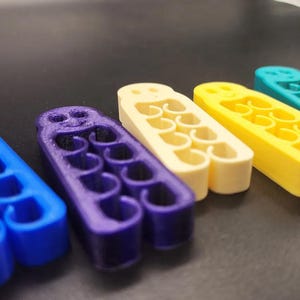 May include: A row of colorful, 3D-printed objects in blue, purple, cream, yellow, teal, and black. Each has a series of curved openings and a rounded top with holes. The objects are arranged on a dark surface.