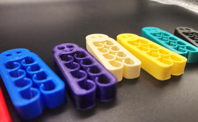 May include: A row of colorful, 3D-printed objects in blue, purple, cream, yellow, teal, and black. Each has a series of curved openings and a rounded top with holes. The objects are arranged on a dark surface.