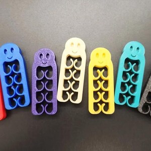 May include: A collection of colorful, 3D-printed cable organizers in red, blue, purple, cream, yellow, teal, and gray. Each organizer has a smiling face and a series of curved slots for holding cables. The organizers are approximately 3 inches long.