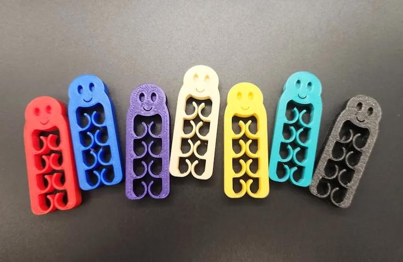 May include: A collection of colorful, 3D-printed cable organizers in red, blue, purple, cream, yellow, teal, and gray. Each organizer has a smiling face and a series of curved slots for holding cables. The organizers are approximately 3 inches long.