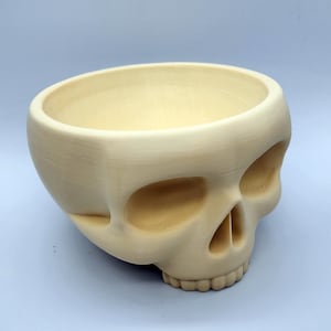 May include: A beige skull-shaped bowl with a wide opening. The bowl has eye sockets, a nose cavity, and a row of teeth. The skull is made of a smooth material and has a slightly rounded shape.
