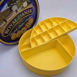 May include: A yellow, round, divided container with a lid, next to a blue tin of Royal Dansk Danish Butter Cookies. The container has multiple compartments for organizing items. The tin has text that reads "Royal Dansk Danish Butter Cookies."