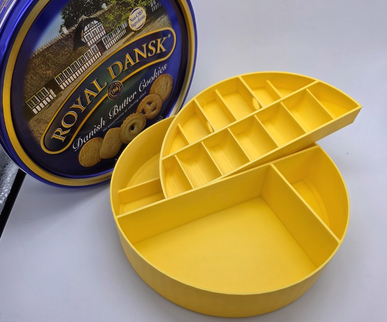 May include: A yellow, round, divided container with a lid, next to a blue tin of Royal Dansk Danish Butter Cookies. The container has multiple compartments for organizing items. The tin has text that reads "Royal Dansk Danish Butter Cookies."