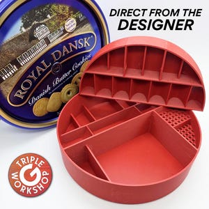 May include: A red, semi-circular organizer with multiple compartments, next to a blue and gold Royal Dansk Danish Butter Cookies tin. The organizer has a grid-like top section and various sized compartments. Text on the tin reads "Direct From The Designer".