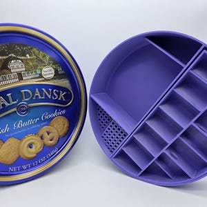 May include: A blue Royal Dansk butter cookies tin next to a purple, circular organizer with multiple compartments. The tin features an image of a Danish village and the words "Danish Butter Cookies". The organizer is designed to hold small items.