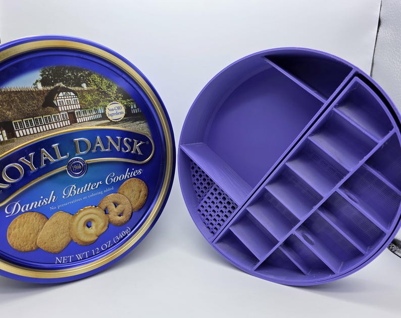 May include: A blue Royal Dansk butter cookies tin next to a purple, circular organizer with multiple compartments. The tin features an image of a Danish village and the words "Danish Butter Cookies". The organizer is designed to hold small items.