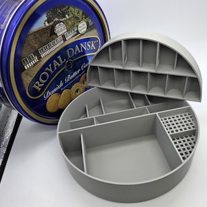 May include: A gray, semi-circular organizer with multiple compartments and a removable lid, next to a blue and gold Royal Dansk Danish Butter Cookies tin. The organizer is designed for storage and organization.