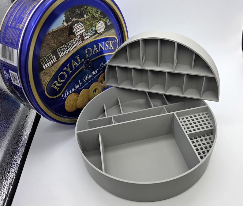 May include: A gray, semi-circular organizer with multiple compartments and a removable lid, next to a blue and gold Royal Dansk Danish Butter Cookies tin. The organizer is designed for storage and organization.