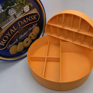 May include: An open orange plastic cookie storage container with a removable lid, next to a blue and gold Royal Dansk Danish Butter Cookies tin. The container has several compartments for organizing cookies. The tin features an image of a building.