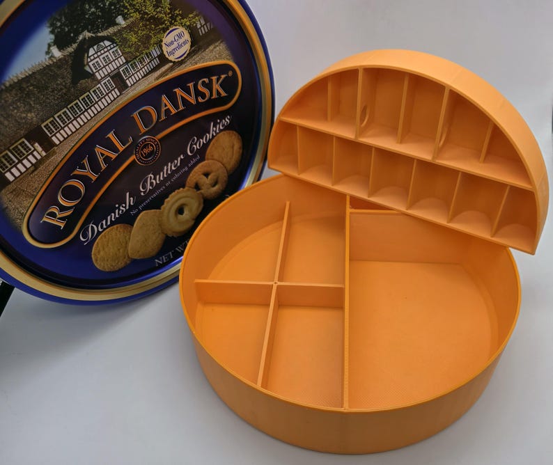 May include: An open orange plastic cookie storage container with a removable lid, next to a blue and gold Royal Dansk Danish Butter Cookies tin. The container has several compartments for organizing cookies. The tin features an image of a building.
