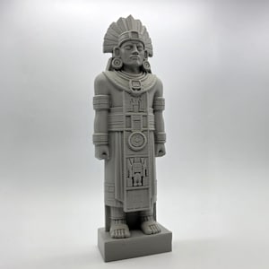 May include: A gray statue of a figure in traditional Aztec attire, including a feathered headdress and ornate clothing. The statue is standing on a rectangular base and has detailed carvings.