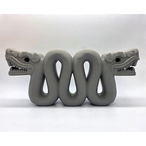 May include: A gray, stylized sculpture of two intertwined serpents, each with detailed heads featuring open mouths and visible teeth. The sculpture has a textured surface and is set against a white background.