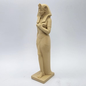 May include: A beige-colored statue of an Egyptian queen, standing on a rectangular base. The queen is adorned with a headdress, armbands, and a draped garment. The statue is a detailed representation of ancient Egyptian art.