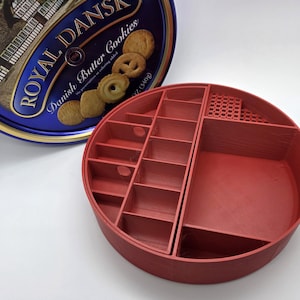 May include: A red, circular organizer with multiple compartments and a blue tin of Royal Dansk Danish Butter Cookies. The organizer has various sized sections for storage. The cookie tin is partially visible, with the words "Royal Dansk" and "Danish Butter Cookies" printed on it.