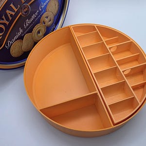 May include: An orange, circular organizer with multiple compartments, designed to fit inside a Royal Dansk Danish Butter Cookies tin. The organizer has various sized rectangular slots and a larger open area.