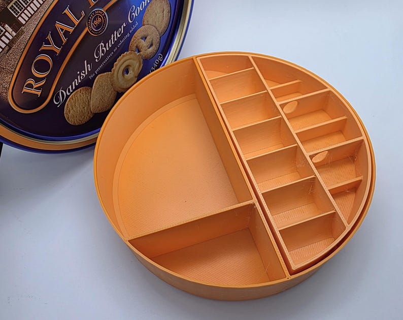 May include: An orange, circular organizer with multiple compartments, designed to fit inside a Royal Dansk Danish Butter Cookies tin. The organizer has various sized rectangular slots and a larger open area.