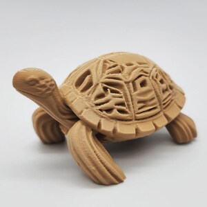 May include: A detailed, tan-colored turtle figurine. The turtle has a textured shell with intricate cut-out designs. Its head and legs are also textured, giving it a realistic appearance. The figurine is set against a plain white background.