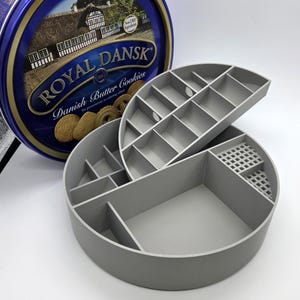 May include: A grey, multi-compartment organiser with a lid, next to a blue and gold Royal Dansk Danish Butter Cookies tin. The organiser has various sized sections for storage. The cookie tin features a picture of a building and cookies.