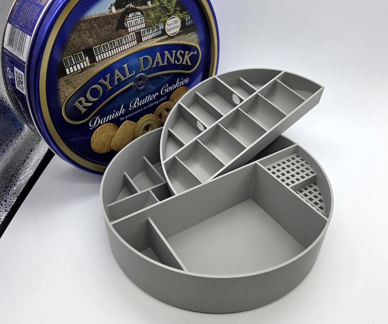 May include: A gray, multi-compartment organizer with a hinged lid, next to a blue and gold Royal Dansk Danish Butter Cookies tin. The organizer has various sized compartments for storage.