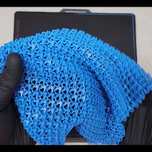 May include: A vibrant blue, flexible, 3D-printed textile. The material is held by a gloved hand, showcasing its intricate, open-weave pattern. The fabric appears to be lightweight and pliable, with a complex, repeating design.