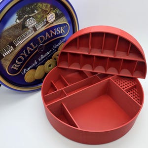 May include: A red, semi-circular, multi-compartment organizer with a lid, next to a blue and gold Royal Dansk Danish Butter Cookies tin. The organizer has various sized compartments and a perforated section. The tin features text and images of cookies.