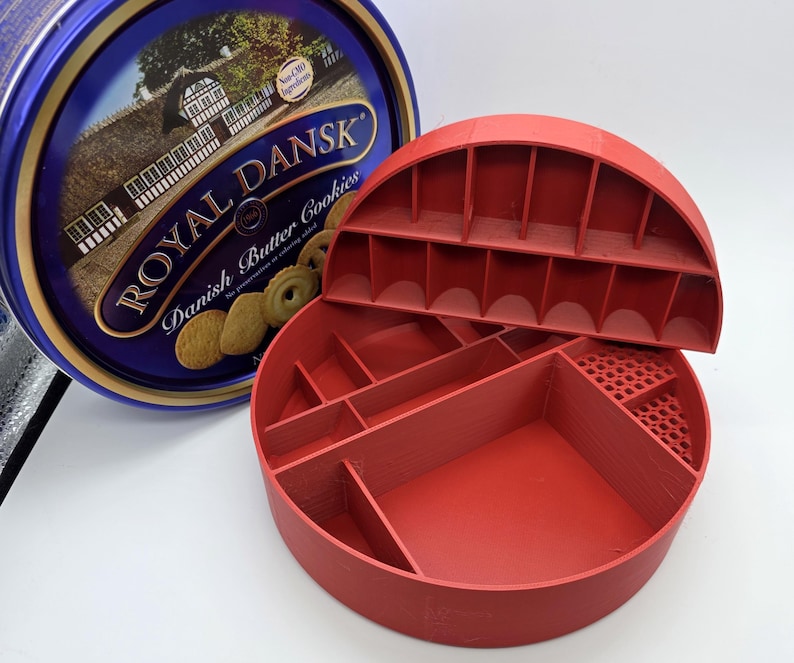 May include: A red, semi-circular, multi-compartment organizer with a lid, next to a blue and gold Royal Dansk Danish Butter Cookies tin. The organizer has various sized compartments and a perforated section. The tin features text and images of cookies.