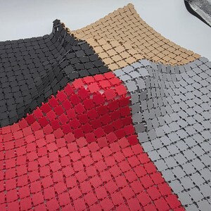 May include: A close-up view of four sections of a flexible, square-tiled material in red, black, tan, and gray. The material is arranged in a wavy pattern, with each tile slightly raised, creating a textured surface. The tiles are uniform in size and shape.