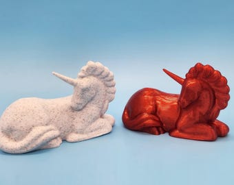 May include: Two unicorn figurines, one white with speckled detailing and the other a vibrant red. Both are in a resting pose, with their horns pointing upwards. The background is a solid light blue.