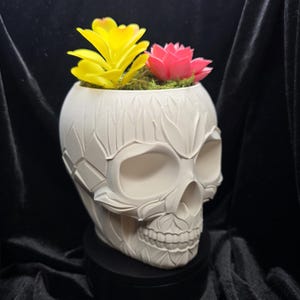 May include: A white skull-shaped planter with carved leaf details, holding yellow and pink artificial flowers and green moss. The planter sits on a black base against a dark background. A unique decorative item.