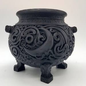 May include: A matte black cauldron featuring intricate relief carvings of crescent moons and swirling cloud patterns. The vessel has a wide rim, two small side handles, and stands on three ornate, carved feet, showcasing a detailed texture.