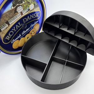 May include: A blue and gold Royal Dansk Danish Butter Cookies tin next to a black, round, divided container. The container has a lid with individual slots. The tin features an image of a castle and cookies.