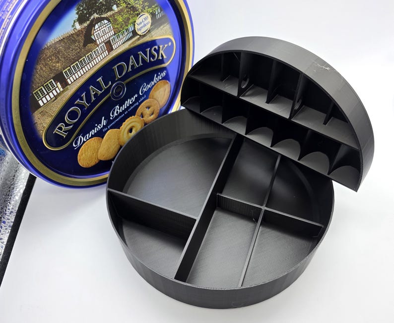 May include: A blue and gold Royal Dansk Danish Butter Cookies tin next to a black, round, divided container. The container has a lid with individual slots. The tin features an image of a castle and cookies.